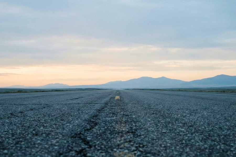 Low-angle wide shot of a textured asphalt highway in Montana disappearing into the horizon at dawn. The sky is overcast with cool blue tones, and subtle mountain silhouettes line the distance. No cars or people are visible.